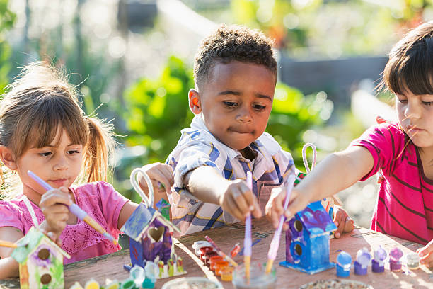 A multiracial group of three serious young children doing an arts and crafts project, painting little wooden bird houses for Earth Day.  It is a bright, sunny day and they are sitting at a wooden table reaching with their paintbrushes.  An African American boy, 5 years old, is sitting in the middle between two Hispanic girls.